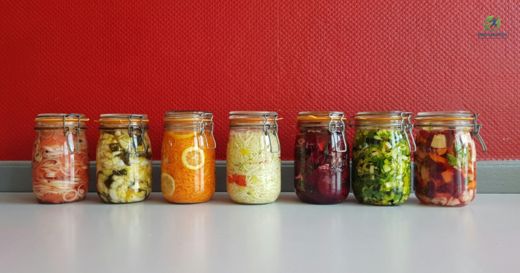 A row of colorful glass jars filled with various fermented vegetables like sauerkraut, kimchi, and pickled lemons against a red background.